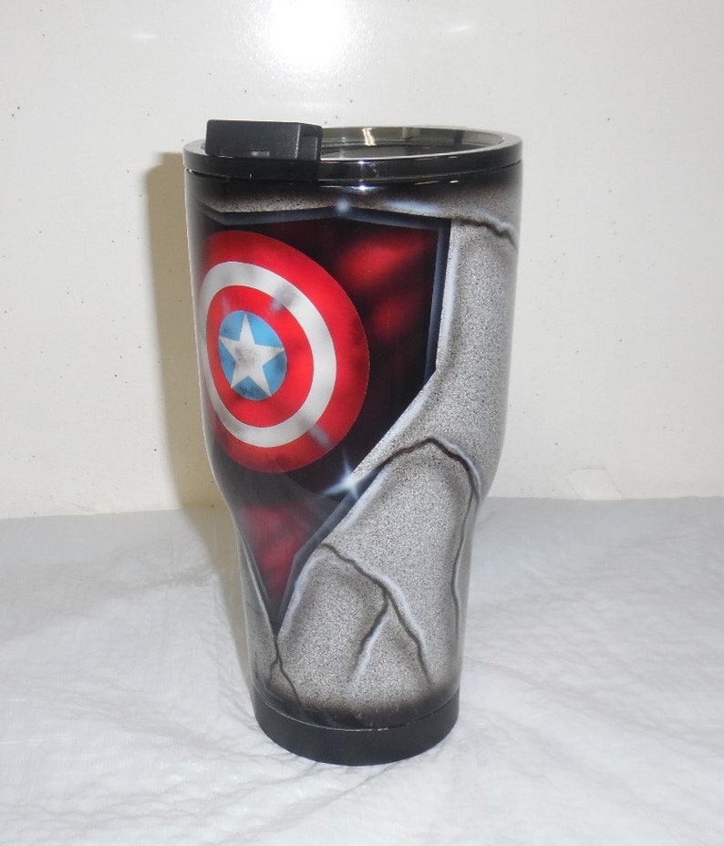 May include: A stainless steel tumbler with a black lid and a graphic of Captain America's shield on a cracked, gray background.