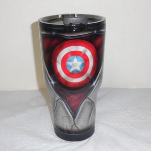 May include: A stainless steel tumbler with a black lid and a custom design featuring Captain America's shield. The shield is red, white, and blue with a white star in the center. The tumbler has a distressed, metallic finish.