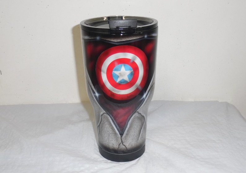 May include: A stainless steel tumbler with a black lid and a custom design featuring Captain America's shield. The shield is red, white, and blue with a white star in the center. The tumbler has a distressed, metallic finish.