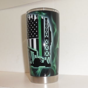 May include: A stainless steel tumbler with a black and green design. The design features an American flag with a green stripe, an oil rig, and an oil pump. The background is a green flame pattern.