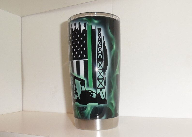 May include: A stainless steel tumbler with a black and green design. The design features an American flag with a green stripe, an oil rig, and an oil pump. The background is a green flame pattern.