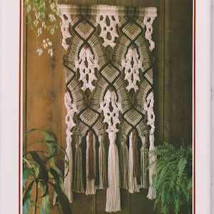 Puede incluir: Un tapiz de macramé para colgar en la pared con un patrón geométrico en tonos marrones, beige y blanco. El tapiz tiene una franja de borlas en marrón y blanco.