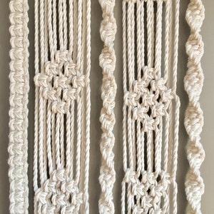 May include: A white macrame wall hanging featuring various patterns and textures. The hanging is crafted from thick cotton rope and has a bohemian aesthetic.