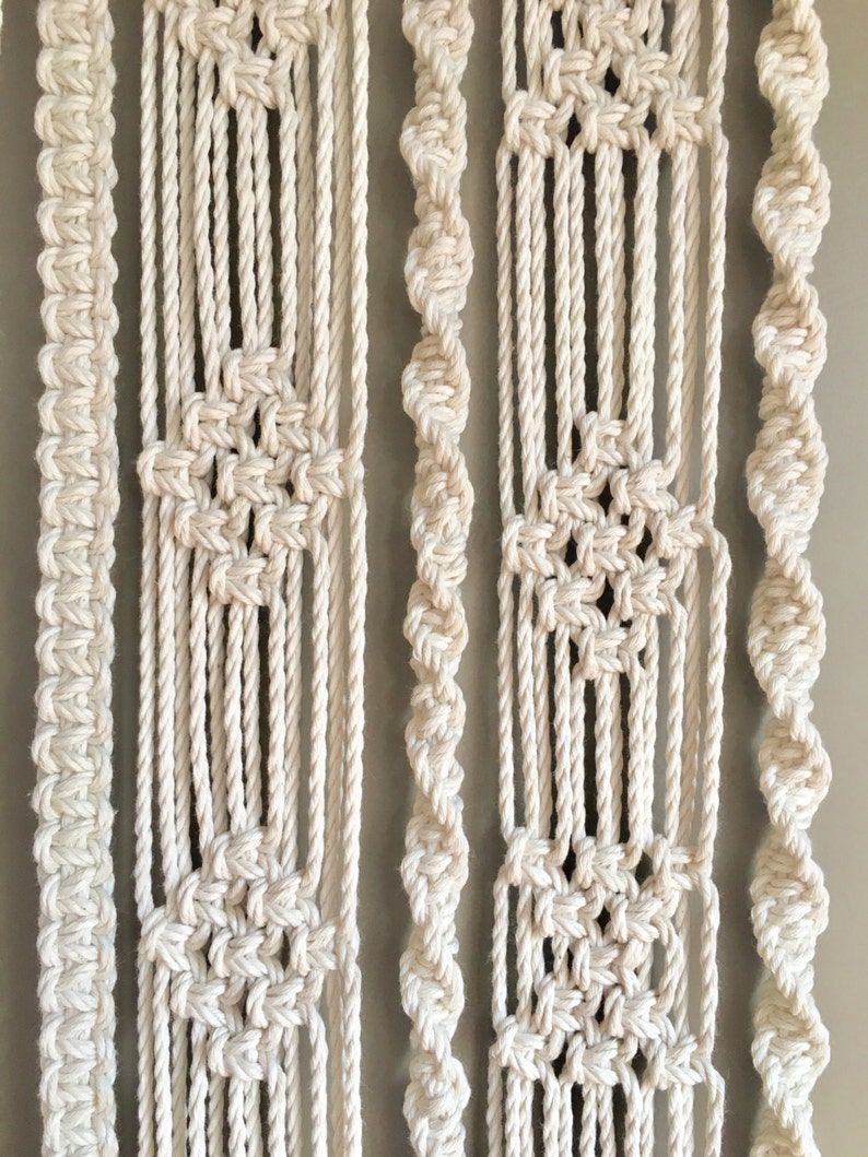 May include: A white macrame wall hanging featuring various patterns and textures. The hanging is crafted from thick cotton rope and has a bohemian aesthetic.