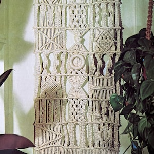 May include: A macrame wall hanging with a geometric pattern in a cream color. The hanging is made of knotted cords and has a fringe at the bottom. The text "Autumn Mixture" is visible at the bottom of the hanging.