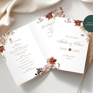 May include: A folded white wedding program card featuring a floral and autumn leaf border in shades of brown and cream. The text reads Welcome to the Wedding of Amber & Oliver, Saturday 11.10.2027, St. Mary Church, Cambridge, and The ceremony.