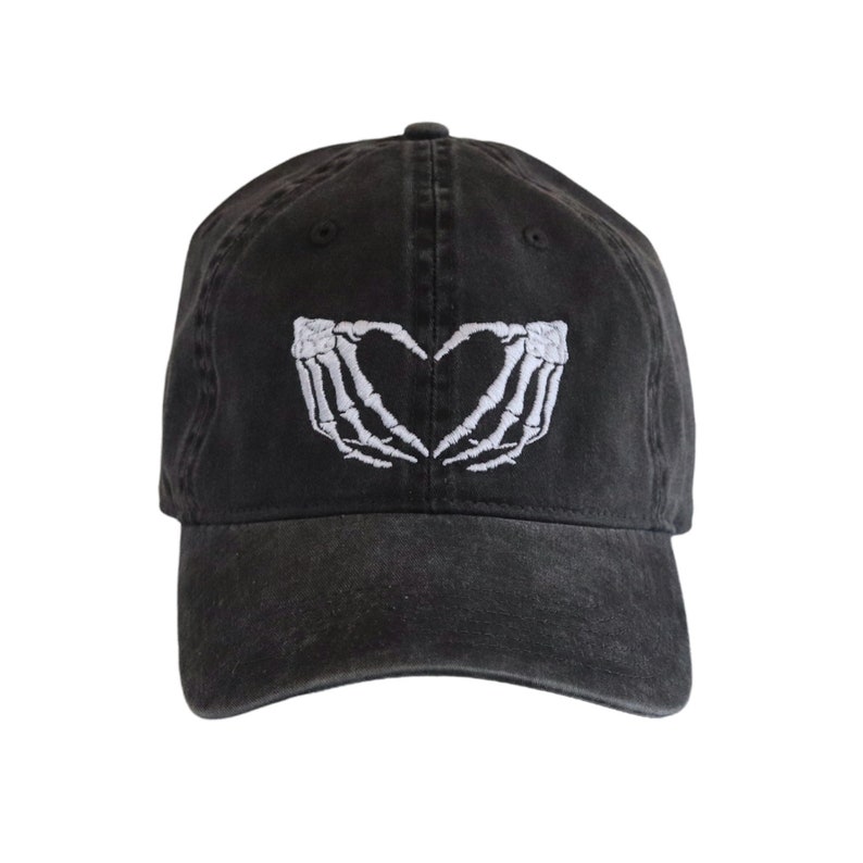 May include: A black baseball cap featuring an embroidered white graphic on the front panel. The design depicts two skeletal hands positioned to form a heart shape. The cap has a curved brim and a structured crown with a button at the top center.