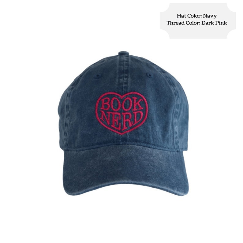 May include: Navy blue baseball cap with the embroidered words "BOOK NERD" inside a heart shape in dark pink thread. The hat is a classic six-panel design with a curved brim.