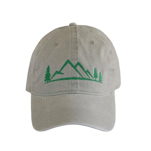 May include: A light gray baseball cap featuring an embroidered green graphic of a mountain range with pine trees on either side. The design is centered on the front panel of the cap, which has a curved brim and a structured crown with eyelets.