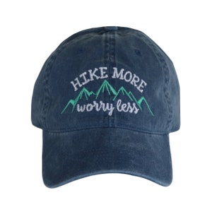 May include: A navy blue baseball cap with an embroidered design featuring a green mountain range and the white text "HIKE MORE" above the mountains and "worry less" below the mountains in a cursive font on the front panel.