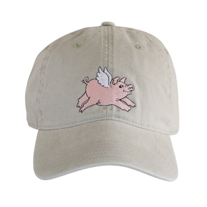 May include: A beige baseball cap featuring an embroidered graphic of a pink pig with white wings. The pig is depicted in a running pose with its legs extended and a small curly tail. The cap has a structured crown, a curved brim, and eyelets.