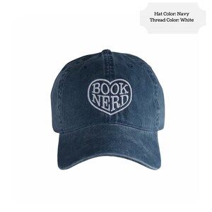 May include: Navy blue baseball cap with the embroidered words "BOOK NERD" inside a white heart-shaped design. The hat is made of a soft material and has a curved brim.