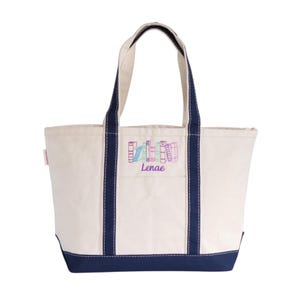 May include: A cream-colored tote bag with navy blue accents. The bag features a small front pocket and navy blue handles. Embroidered on the front is a stack of colorful books with the name "Lenae" in purple script.