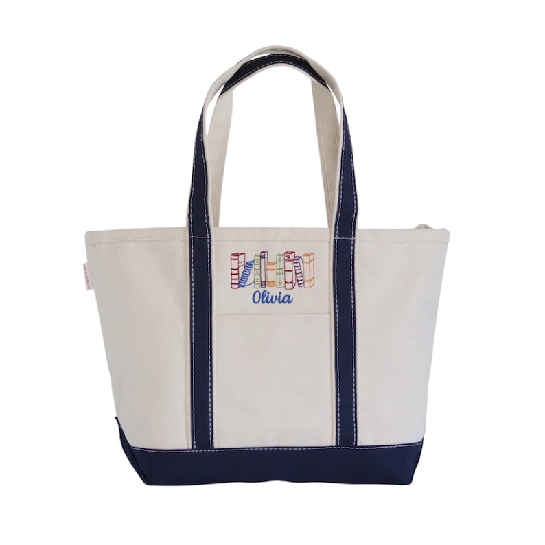 May include: Cream-colored canvas tote bag with navy blue trim and handles. The bag features an embroidered design of colorful books and the name "Olivia" in blue. The tote has a front pocket and a zipper closure.
