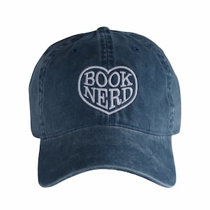 May include: A navy blue baseball cap with a curved brim. The cap features the embroidered words "BOOK NERD" inside a white heart shape. The hat is made of a soft, worn material.