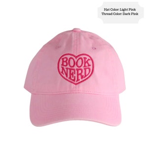 Light pink baseball cap embroidered with the words "Book Nerd" inside a heart shape using dark pink embroidery thread.