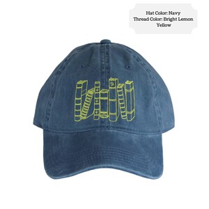 May include: A navy blue baseball cap with a yellow embroidered design of a stack of books. The text "Hat Color: Navy Thread Color: Bright Lemon Yellow" is printed on a white rectangle above the cap.