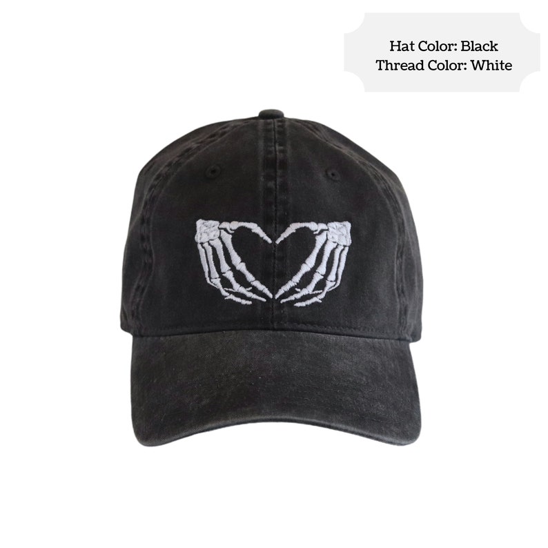 May include: A black baseball cap featuring white embroidery of two skeleton hands forming a heart shape on the front panel. The text on the image reads: Hat Color: Black, Thread Color: White. The cap has a curved brim and a structured crown.
