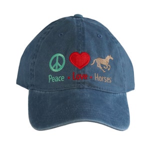 May include: A navy blue baseball cap featuring embroidered designs on the front: a light green peace symbol, a red heart, and a tan horse. Below these icons, the text reads Peace, Love, and Horses in matching colors, centered on the crown.
