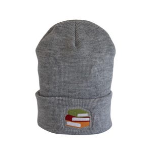 May include: A gray beanie with a folded brim. The beanie features an embroidered design of a stack of books in green, red, and orange. The hat is made of a soft, knitted material, perfect for cold weather.