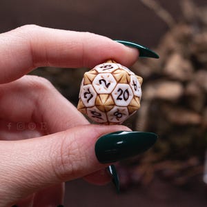 Könnte beinhalten: Ein zwanzigseitiger Würfel mit einem geometrischen Design in Weiß und Hellbraun sowie schwarzen Ziffern. Der Würfel hat eine strukturierte, mehrflächige Oberfläche mit erhabenen Kanten. Der Text RESIN CAST REPLICAS CRUELTY FREE ist rechts sichtbar.
