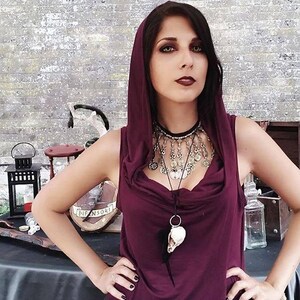 A woman is wearing a garnet hoodie with a silver and black necklace. The necklace has a skull pendant. The woman stands in front of a wooden table with a lantern and other objects.