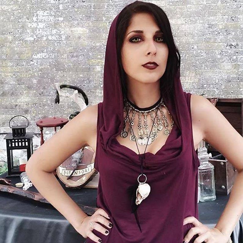 A woman is wearing a garnet hoodie with a silver and black necklace. The necklace has a skull pendant. The woman stands in front of a wooden table with a lantern and other objects.