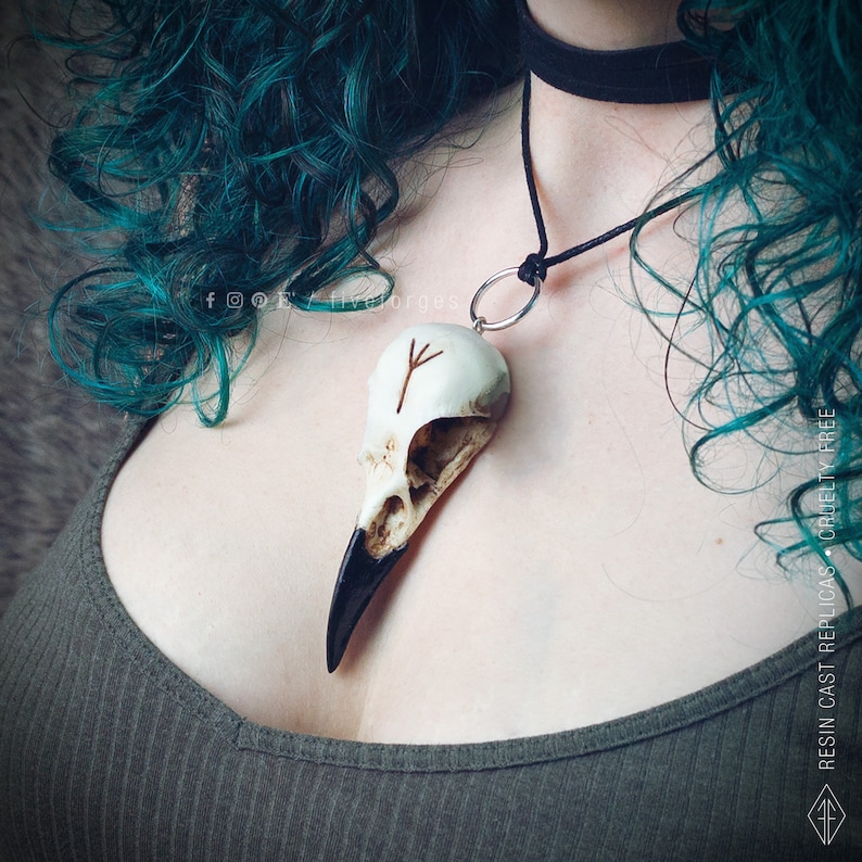 Realistic crow skull resin replica with an engraved brown Elder Futhark rune, with a black cord necklace and a silver ring, worn by a girl. Customizable norse rune.