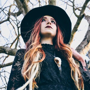 A woman with long red and blonde hair wears a wide-brimmed black hat and a black lace dress. She wears a necklace with a crow skull pendant. The background is a blurry image of a tree.