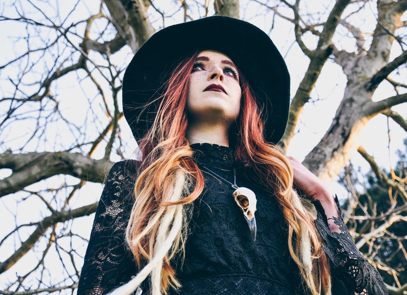 A woman with long red and blonde hair wears a wide-brimmed black hat and a black lace dress. She wears a necklace with a crow skull pendant. The background is a blurry image of a tree.
