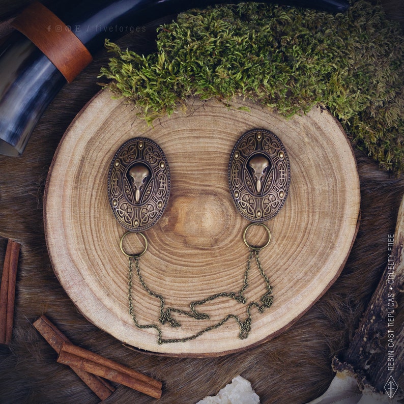 Two hand painted bronze brooches with a bird crow skull design, connected by two bronze chains. The pins are on a wooden surface. Perfect for historical reenactments, cosplay, Viking and Norse style costumes. Medieval wedding dress.