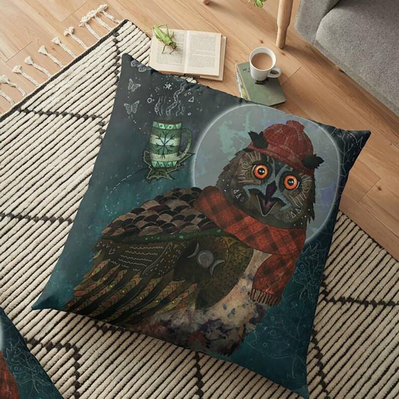 May include: A square floor pillow featuring a whimsical illustration of an owl wearing a red hat and scarf. The owl has large orange eyes and is set against a dark blue background with a full moon and a floating mug of tea. The pillow is likely for home decor.