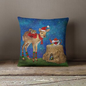 May include: Decorative square pillow featuring a whimsical illustration. A fawn wearing a Santa hat and scarf stands near a tree stump with a teapot. Two squirrels ride on the fawn's back. The background is a starry night sky.