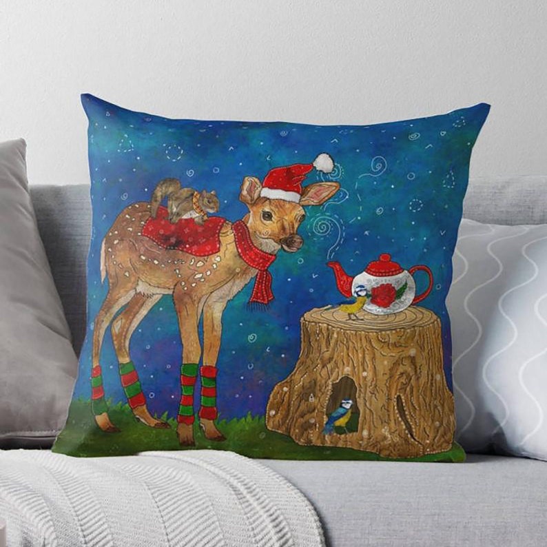 May include: Decorative throw pillow featuring a whimsical winter scene. A deer wearing a Santa hat and scarf stands near a tree stump with a teapot. A squirrel rides on the deer's back. The background is a dark blue with white dots.
