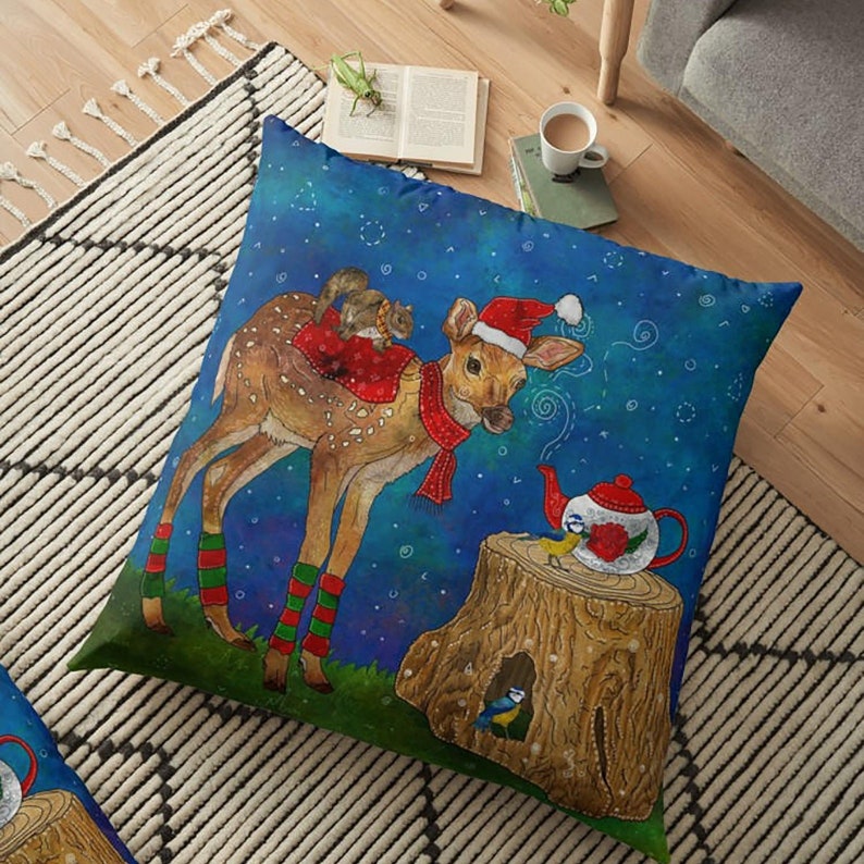 May include: Square floor pillow with a whimsical illustration of a deer wearing a Santa hat and scarf, with a squirrel on its back. A teapot and a tree stump with a bird are also featured. The background is a starry blue.
