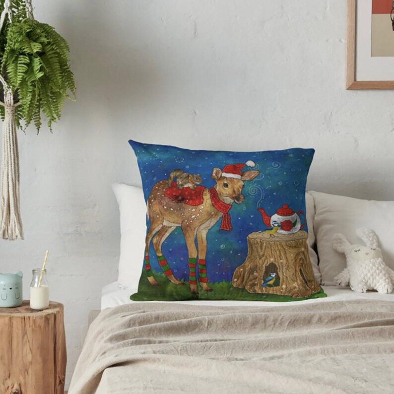 May include: Square decorative pillow with a whimsical illustration of a deer wearing a Santa hat and scarf, with a squirrel on its back. A teapot and birds are on a tree stump against a starry blue background. The pillow is on a bed.