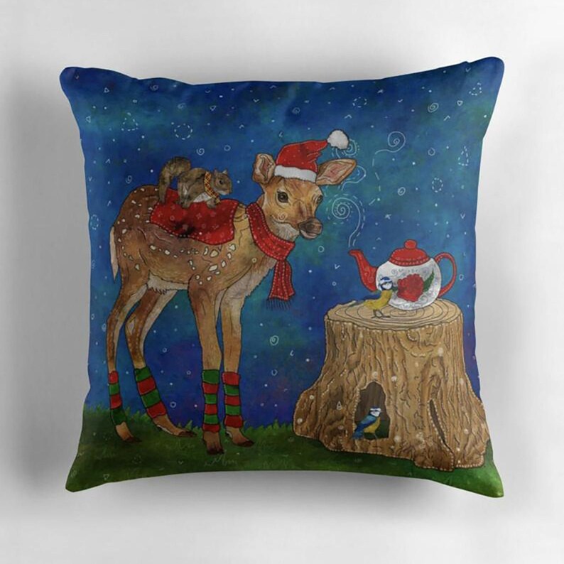 May include: Decorative throw pillow with a whimsical Christmas scene. A deer wearing a Santa hat and scarf has a squirrel on its back. A teapot sits on a tree stump with a bird. The background is a dark blue with white stars.