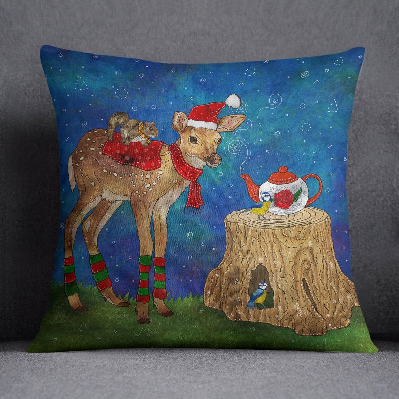 May include: Decorative square pillow featuring a whimsical illustration. A fawn wearing a Santa hat and scarf stands next to a tree stump with a teapot and birds. A squirrel rides on the fawn's back. The background is a starry night sky.