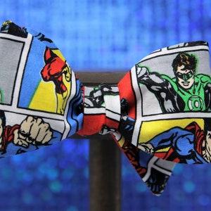 Puede incluir: Una pajarita con un patrón de cómic con Superman, Linterna Verde y otros superhéroes. La pajarita está hecha de tela y tiene una combinación de colores rojo, azul, amarillo y verde.