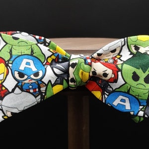 May include: A white bow tie with a colorful pattern of superhero characters, including Iron Man, Captain America, Hulk, and Thor. The bow tie is made of fabric and has a white background.