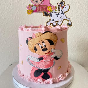 May include: A pink frosted cake decorated with a Minnie Mouse graphic, a number one, and a cartoon horse. The cake has pink frosting and gold leaf accents. The Minnie Mouse graphic is wearing a pink dress and a straw hat. The cake is on a silver cake stand.