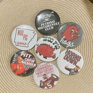 May include: A collection of round buttons with various designs and text. One button features a motorcycle, another says "Woo Pig Sooie" with an outline of Arkansas, and others display sports-related graphics and phrases like "Horns Down."