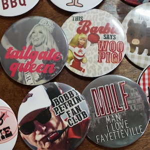May include: A collection of round buttons with various designs and text. One button features a woman with the text "tailgate queen." Another says "This Barbie says Woo Pig!" Other buttons have text like "Bobby Petrino Fan Club" and "MILF Man I Love Fayetteville."