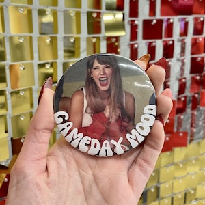 May include: A round button with a photo of a woman with long brown hair, wearing a white top and red clothing, and with her mouth open in a scream. The button has the words "GAMEDAY MOOD" in white, curved around the image.