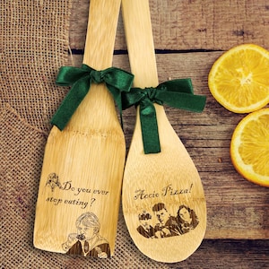 May include: Two wooden kitchen spoons tied with green bows. One spoon has the text "Do you ever stop eating?" and a cartoon image of a person. The other spoon has the text "Accio Pizza!" and a cartoon image of four people.