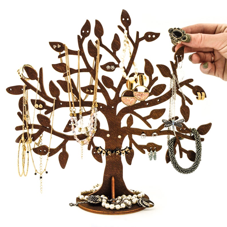 Dark brown wooden jewelry holder. The tree has holes for earrings. Other jewelry can be hung on the branches. Several pairs of earrings, chains, rings and a bracelet are hung on the tree. Next to it is a woman's hand hanging a ring on a tree.