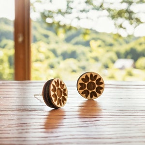May include: A pair of wooden earrings with a carved sunflower design. The earrings are brown and have a natural wood finish.