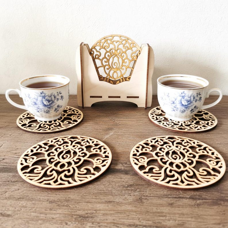 4 wooden coasters are placed on the table. Tea cups are placed on 2 coasters. There is a coaster holder with coasters inside. Placemats are cut out with floral motifs.