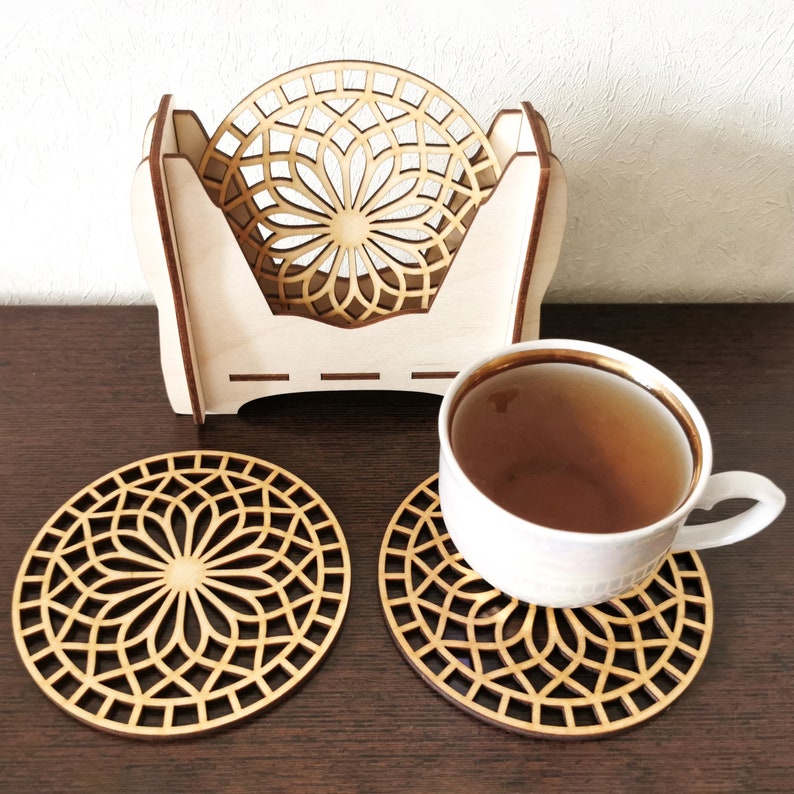 2 wooden coasters are placed on the table. A cup of tea is placed on 1 coaster. There is a coaster holder with coasters inside. Placemats are cut out with modern flower motifs.