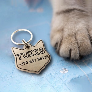 May include: Silver cat ID tag with a black outline and the name "TUKIS" and phone number +370 657 88135 engraved on it. The tag is attached to a silver key ring.